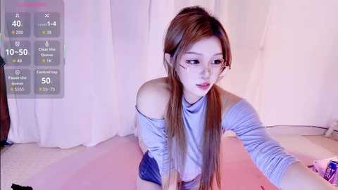 Media: A young East Asian woman with long brown hair, wearing a light gray off-shoulder top and denim shorts, is leaning forward, reaching out with a selfie stick, in a softly lit, pink room with a white curtain.