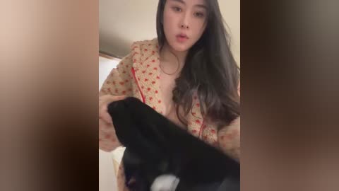 Video of an Asian woman with long, straight black hair, wearing a beige floral robe, holding a black jacket in a dimly lit room with a blurred background.