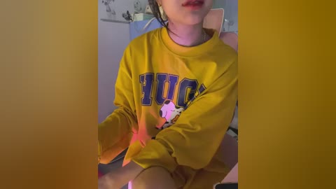 Video of a young woman with light skin and long dark hair, wearing a mustard-yellow oversized \"PIKE\" sweatshirt and sitting on a chair, illuminated by a purple light.
