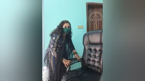 Video of a woman in a black, gold-patterned shalwar kameez, wearing a green mask, holding a book, standing in a teal-painted room with a black leather chair and wooden door.