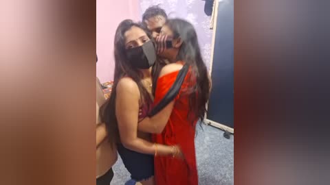 Video of three South Asian women in a dimly lit room, one wearing a red sari, another a black mask, embracing, with a blurred person in the background.