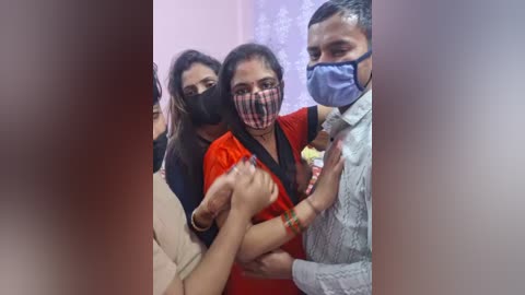 Video of three people wearing face masks and traditional attire, holding hands, in a crowded indoor setting with blurred background.