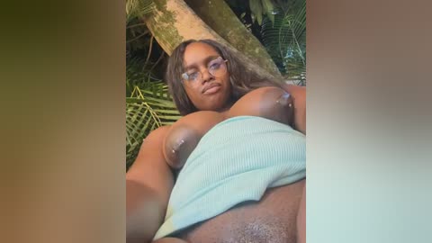 Media: A video of a Black woman with long, wavy brown hair, wearing glasses and a light blue crop top, partially exposed breasts, and lying on a lush tropical forest floor.