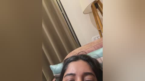 A close-up video of a person with long dark hair, partially visible face, lying on a bed with beige and light blue bedding. A wooden lamp and beige curtain are in the background.