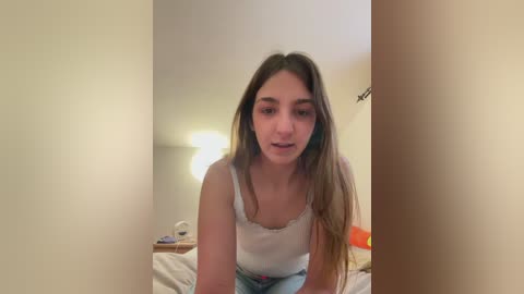 Video of a young woman with long, straight brown hair, wearing a white tank top and light blue jeans, sitting on a bed in a dimly lit room with cream-colored walls.