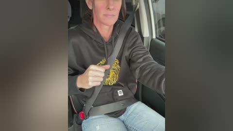 Video of a middle-aged woman with light skin and dark hair wearing a black hoodie with a yellow tiger graphic, driving a car with a gray seatbelt.