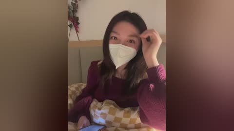Video of an Asian woman with long black hair, wearing a white face mask, maroon sweater, and checkered blanket, sitting in a dimly lit room with a beige wall and a wooden headboard.