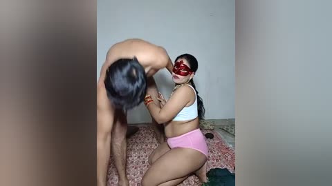 Video of a shirtless man and a woman in a red mask and pink shorts kneeling on a patterned rug, with a plain wall background.