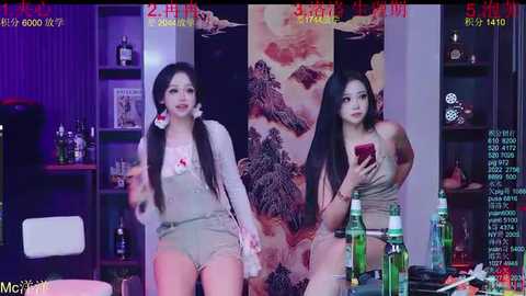 Video of two East Asian women with long black hair, one in overalls, the other in a dress, holding a red phone, standing in a dimly lit room with alcohol bottles and a green bottle.