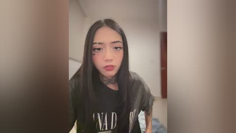 Video of a young woman with long black hair, wearing a black shirt with \"MAD\" in white letters, standing in a dimly lit room with blurred background.