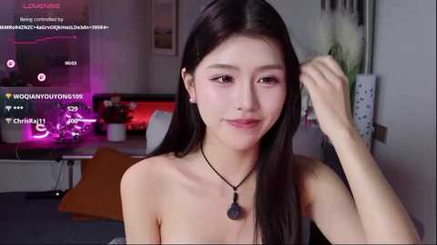 Video of an East Asian woman with long black hair, fair skin, and a slender physique, wearing a strapless black top, in a cozy indoor setting with neon lights and a white wall.