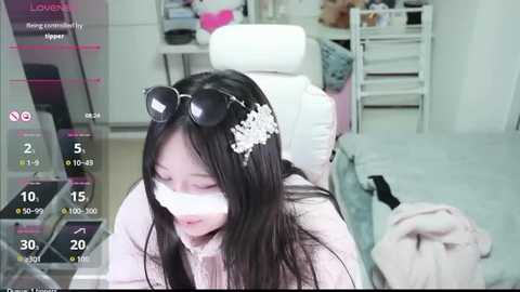 A video of an Asian woman with long black hair, wearing a floral hair accessory, a white face mask, and a pink shirt, lying on a bed with a white chair in the background.