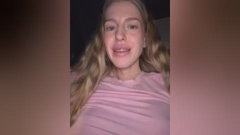 Video of a young woman with long, wavy blonde hair and fair skin, wearing a light pink t-shirt. She has braces and is smiling. The background is dark and blurred.