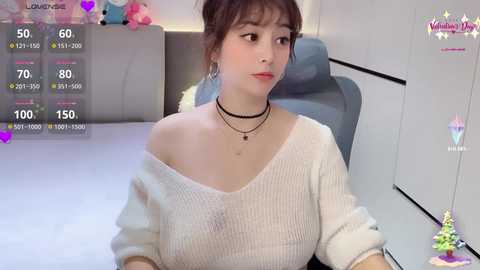 Video of a young Asian woman with fair skin, wearing a white off-shoulder sweater, black choker, and dangling earrings, sitting on a gray chair. Background includes a gray couch and a toy shelf.