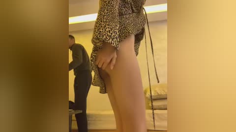 Video of a woman lifting a leopard-print skirt, revealing bare buttocks, in a dimly lit, modern room with beige walls and a bed in the background.