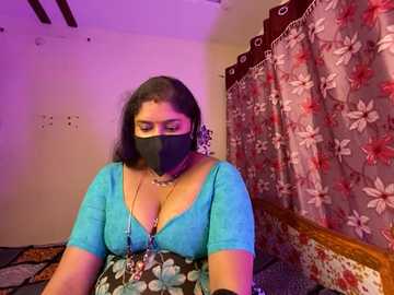 Media: Video of a South Asian woman with medium skin tone and long dark hair, wearing a turquoise blouse and floral-patterned skirt, black mask, and beaded necklace. Background features floral curtains and a wooden headboard.