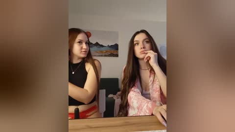 Video of two young women, one in a black crop top, the other in a floral robe, sitting at a wooden table, with a blurred background.