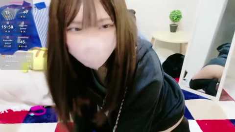 Video of an Asian woman with long brown hair, wearing a black hoodie and a pink face mask, bending over in a bedroom with white walls, a round table, and a green plant.