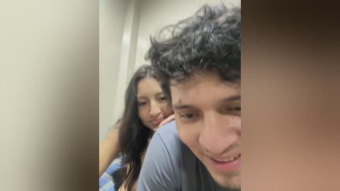 Video of a young woman with long black hair and a man with curly dark hair, both smiling, in a narrow corridor with beige walls.