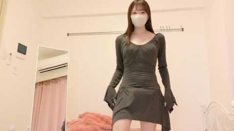 Video of a slender East Asian woman in a gray dress, mask, and gloves, standing in a beige room with a bed, air conditioner, and curtain.