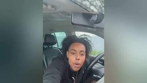 A video of a young Black man with dreadlocks, wearing a black hoodie, driving a car with a blurred background, and an out-of-focus, blurred image on the right side.