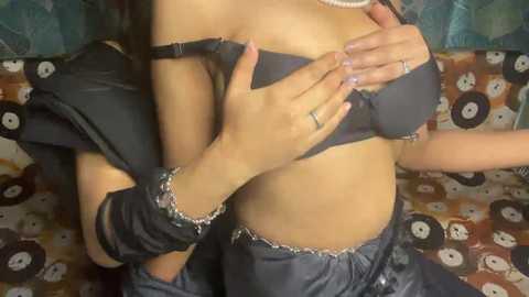 Video of a woman in a black bra, removing a black shirt, revealing her small breasts, adorned with a silver bracelet. Background features a colorful, patterned blanket.