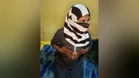A video of an elderly woman wearing a black and white striped hijab, seated on a blue and green patterned blanket against a mustard-yellow wall. She appears to be in a domestic setting.
