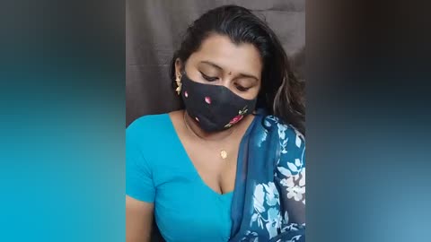 Video of a South Asian woman with medium-brown skin, wearing a blue floral sari, black mask, and earrings, with a blurred background.