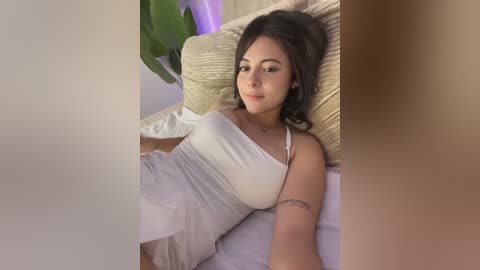 Video of a young woman with light brown skin, long dark hair, and medium-sized breasts, lying on a beige pillow in a cozy, dimly lit room, wearing a white tank top and light-colored shorts.