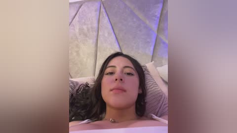 A video of a young woman with long dark hair, fair skin, and light makeup, lying on a bed with a textured, geometric headboard. She wears a light-colored top. The room is softly lit with purple ambient lighting.