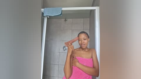 A video of a young Black woman with medium-brown skin, in a bathroom, holding a long, erect penis with her right hand while wrapped in a pink towel, standing in front of a white-framed mirror.