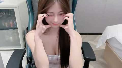 Video of an East Asian woman with long brown hair, wearing a gray tank top, sitting in a blue gaming chair, forming a heart shape with her fingers on her face, in a modern room with beige walls and a bed.