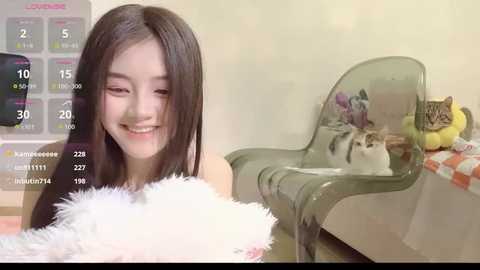 Video of a smiling East Asian woman with long black hair, wearing a white fluffy blanket, sitting on a transparent chair with a cat in her lap, in a brightly lit room.