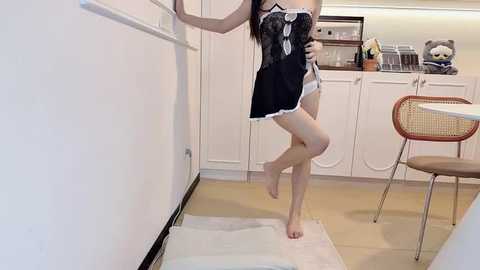 Media: Video of an Asian woman with long black hair, wearing a black lace dress with white trim, standing barefoot in a white kitchen, holding a phone.