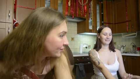 Video of two young women in a kitchen, one with long brown hair in a red top, the other with short brown hair in a white tank top, smiling. Wooden cabinets, hanging red pot holders, and a tiled backsplash.