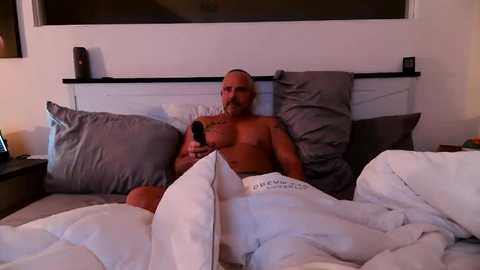 Video of a shirtless, middle-aged, muscular man with a beard, holding a remote, partially covered by white sheets on a bed with gray pillows and a black headboard.