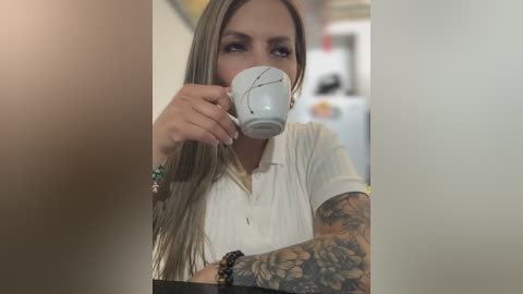 Media: Video of a tattooed woman with long blonde hair, wearing a white blouse, drinking from a white mug, with a blurred background.