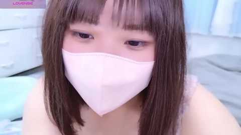 A close-up video of an East Asian woman with straight, dark brown hair and bangs, wearing a light pink surgical mask and a white tank top. The background shows a light blue curtain and a bed.