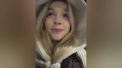 A video of a young, fair-skinned girl with long blonde hair, wearing a hooded gray cloak, smiling gently. The background is blurry, focusing attention on her serene expression.