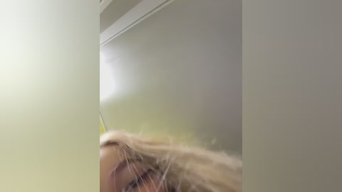 A close-up video shows a blonde woman's face partially obscured by a shiny, reflective surface, possibly a mirror or a glass door, creating a distorted, blurred reflection. The background is a soft, muted green, and her hair appears to be blowing in the wind.