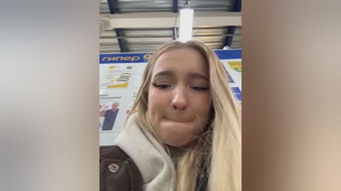 Video of a blonde woman with closed eyes, pouty lips, and a white coat, in a brightly lit room with posters and a blue ceiling.