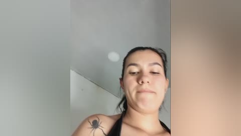 Video of a young woman with light brown skin, dark hair pulled back, and a black spider tattoo on her shoulder, smiling slightly. She's wearing a black top. Background includes a white wall and a beige wall.