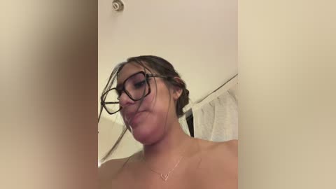 Video of a young woman with light skin, wearing glasses, a wet, messy bun, and no shirt, standing in a shower with a white curtain in the background.