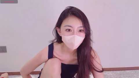 Video of an East Asian woman with long black hair, wearing a blue dress, white mask, and sitting on a gray carpet in a plain room with a beige wall.