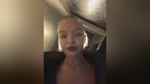 A video of a young, fair-skinned woman with a straight, blonde ponytail, wearing dark lipstick and a black leather jacket. She's indoors with dim lighting, and a chandelier is visible in the background.