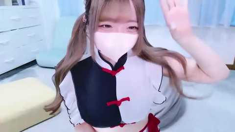 Video of a young, fair-skinned Asian woman with long, light brown hair, wearing a white and black cosplay outfit, mask, and a yellow mat, indoors.