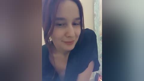 Video of a smiling young woman with shoulder-length auburn hair, wearing a black blouse, in a dimly lit room with a blurred background.