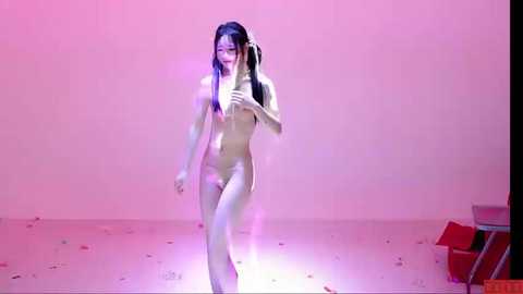 Video of a nude Asian woman with long black hair, standing on a stage with a pink backdrop. She has a slender, athletic build and is illuminated by soft lighting.