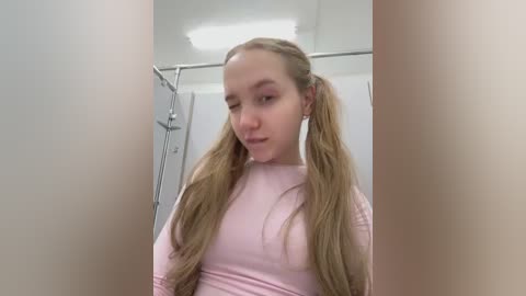 Video of a young woman with long, blonde pigtails, wearing a light pink top, standing in a bathroom with white walls and a silver shower curtain.