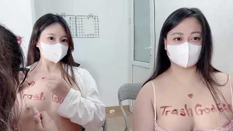 Video of a young Asian woman with long black hair, wearing a white mask and pink bra, standing in a plain room with a grid wall and a chair.